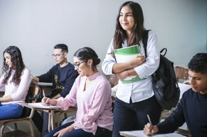 Students seated in a bright classroom while one stands holding a notebook